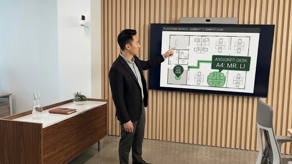 A colleague in a navy jacket points at a lobby display showing a floor plan with a You are here pin and Assigned desk A4: Mr. Li highlighted in green.