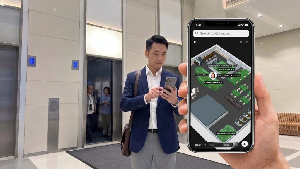 In a lift lobby, a colleague checks his phone while a hand holds a phone overlay searching for a colleague on a 3D floor map, with Sarah J. found in Conference Room B.
