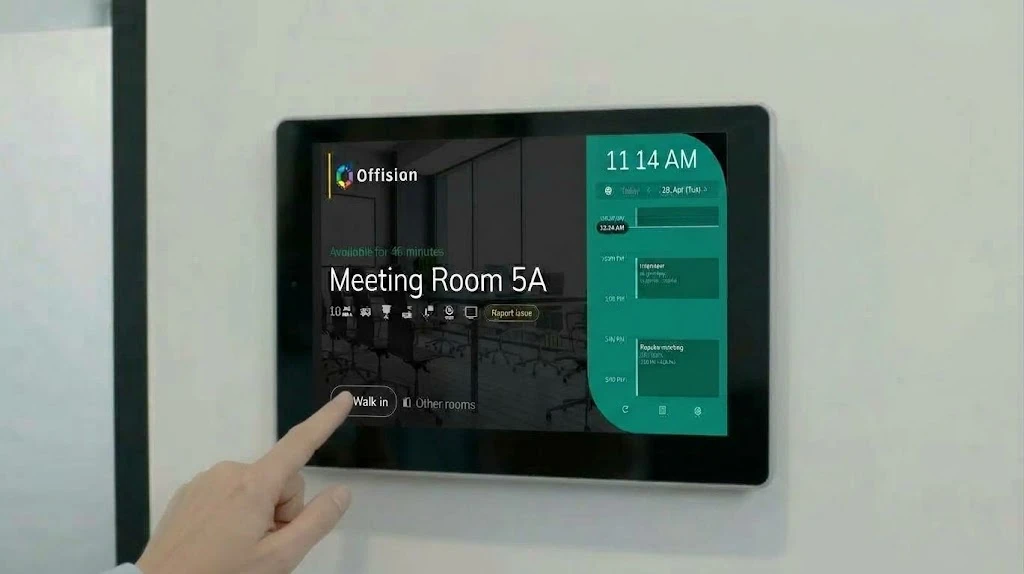A finger taps Walk in on a wall panel showing Meeting Room 5A is free for 46 minutes, with today's schedule beside it.