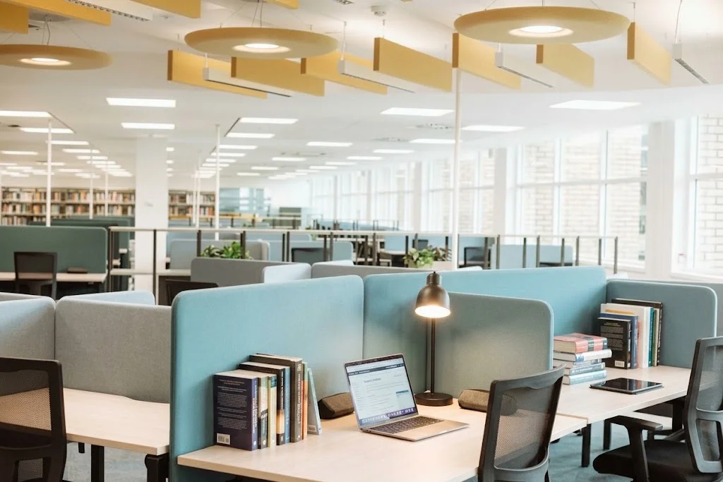A bright library study area with blue-partitioned desks, open laptops, textbooks, and bookshelves in the background